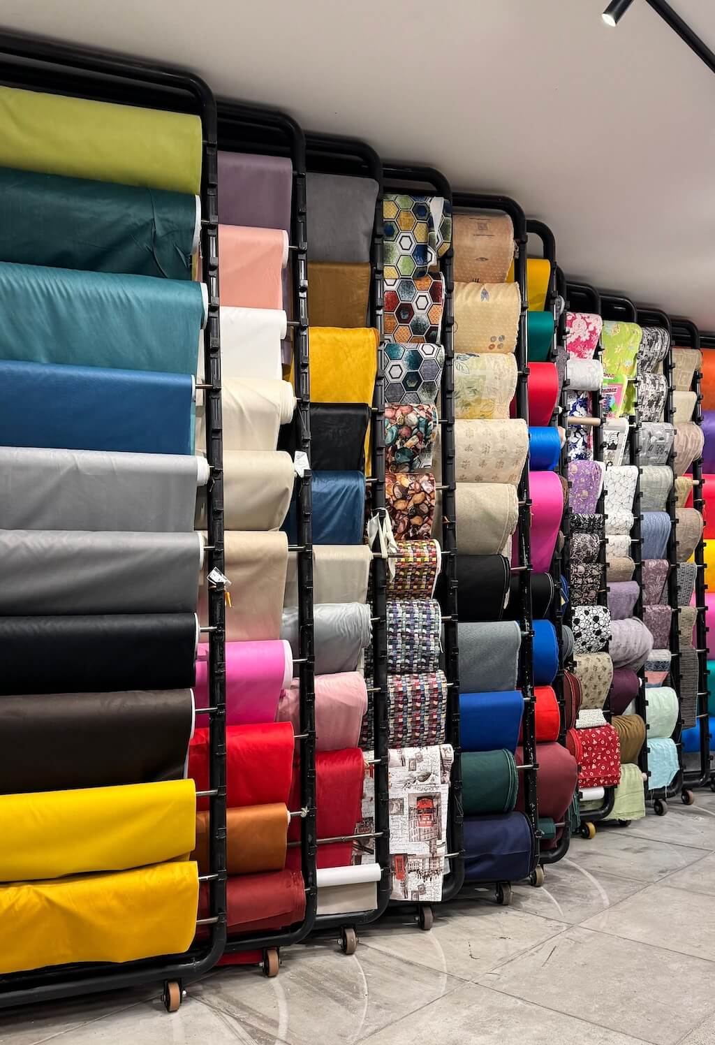 Interior da loja Kami Tecidos no Brás - Ampla variedade de tecidos sintéticos organizados em expositores, mostrando rolos de tecidos em diversas cores e padrões